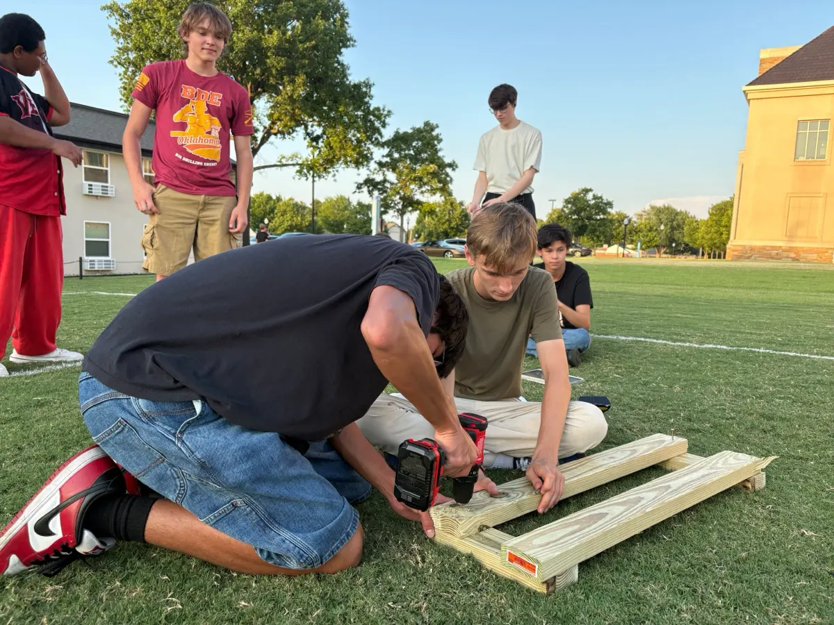 Four TASC students kneeling on a campus lawn at golden hour, drilling together the wooden frame of a trebuchet while two others watch.